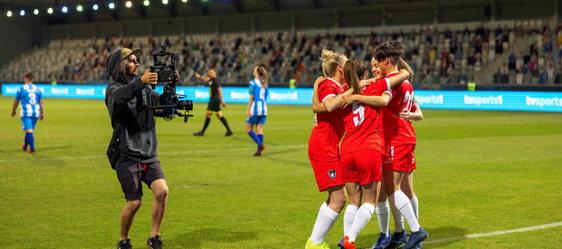 This is a photo of a group of female footballers celebrating, while a man with a camera follows them, filming for the TV